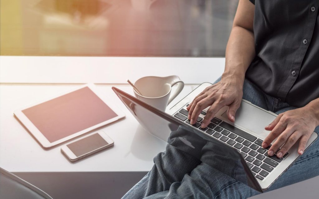 Man works on a laptop while sitting next to a mug, cell phone, and electronic tablet